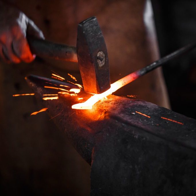 The blacksmith manually forging the molten metal on the anvil in smithy with spark fireworks